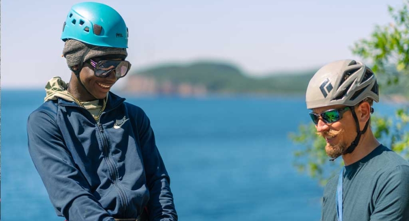 Two people wearing helmets smile. They appear to be at a high elevation. There is water below them.